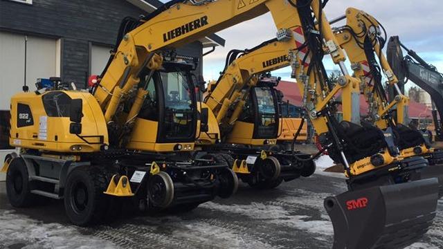 Tre gule Liebherr gravemaskiner parkert foran en bygning, klare for arbeid i snøen.
