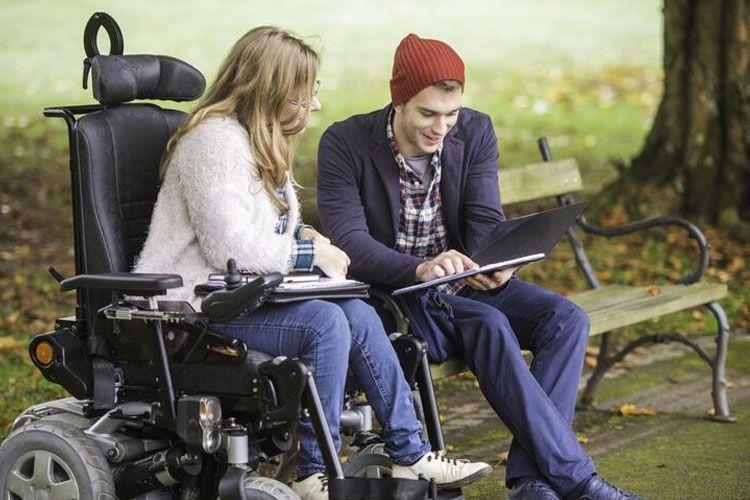 To personer, en kvinne i rullestol og en mann, sitter på en benk og ser på en laptop i en park.