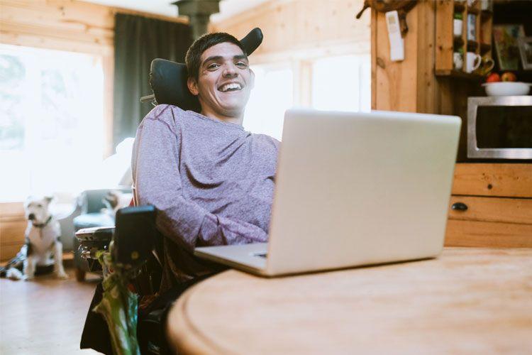 En smilende mann i rullestol sitter ved et bord med en laptop i en koselig hytte.