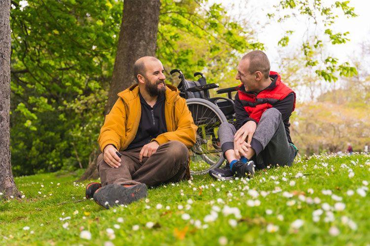 To personer som sitter på gresset i en park, med en rullestol i bakgrunnen og trær rundt dem.