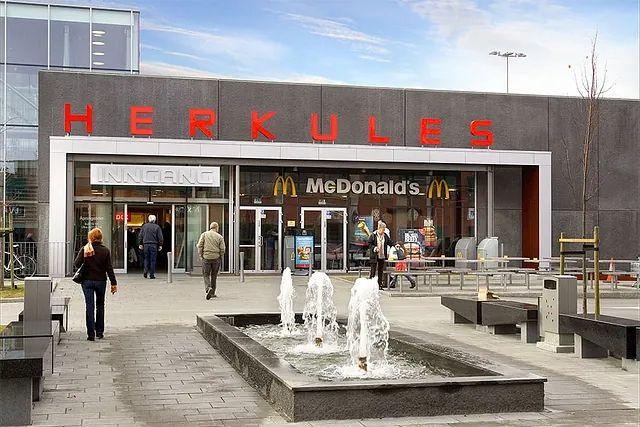 Inngangen til Herkules kjøpesenter med McDonald's og fontener foran. Besøkende går inn i senteret.
