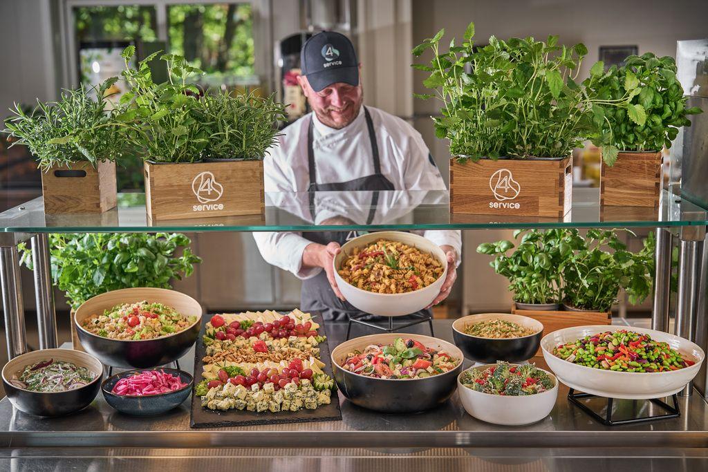 Kokk som serverer en stor bolle med pasta foran et utvalg av friske salater og urter i en restaurant.
