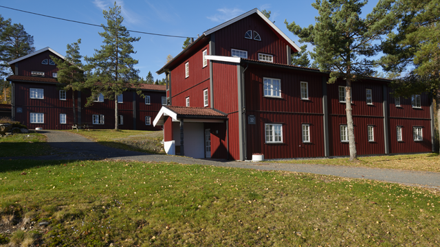 Rødt trehus med flere etasjer, omgitt av grønne trær og en grusvei i solfylt vær.