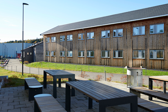 Uteområde med benker og bord foran en trebygning med vinduer og gressplen.