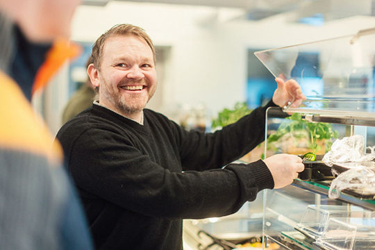 En smilende mann i svart genser som serverer mat fra en glassdisk i en kafé.