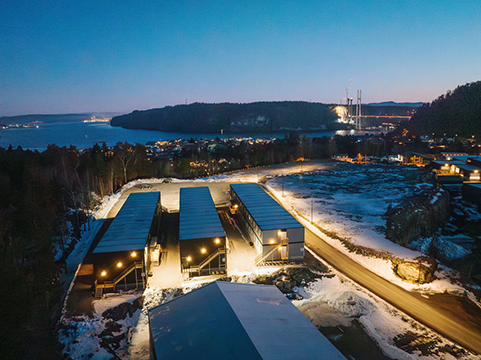 Utsikt over et industriområde med bygninger og snødekte veier ved sjøen i skumringen.