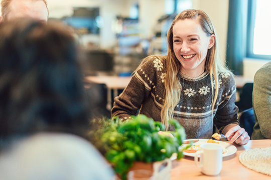 En kvinne med langt hår smiler mens hun spiser frokost sammen med venner i en koselig kafé.