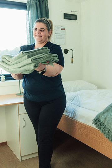 En kvinne holder en stabel med håndklær mens hun står ved en seng i et rom med vindu.