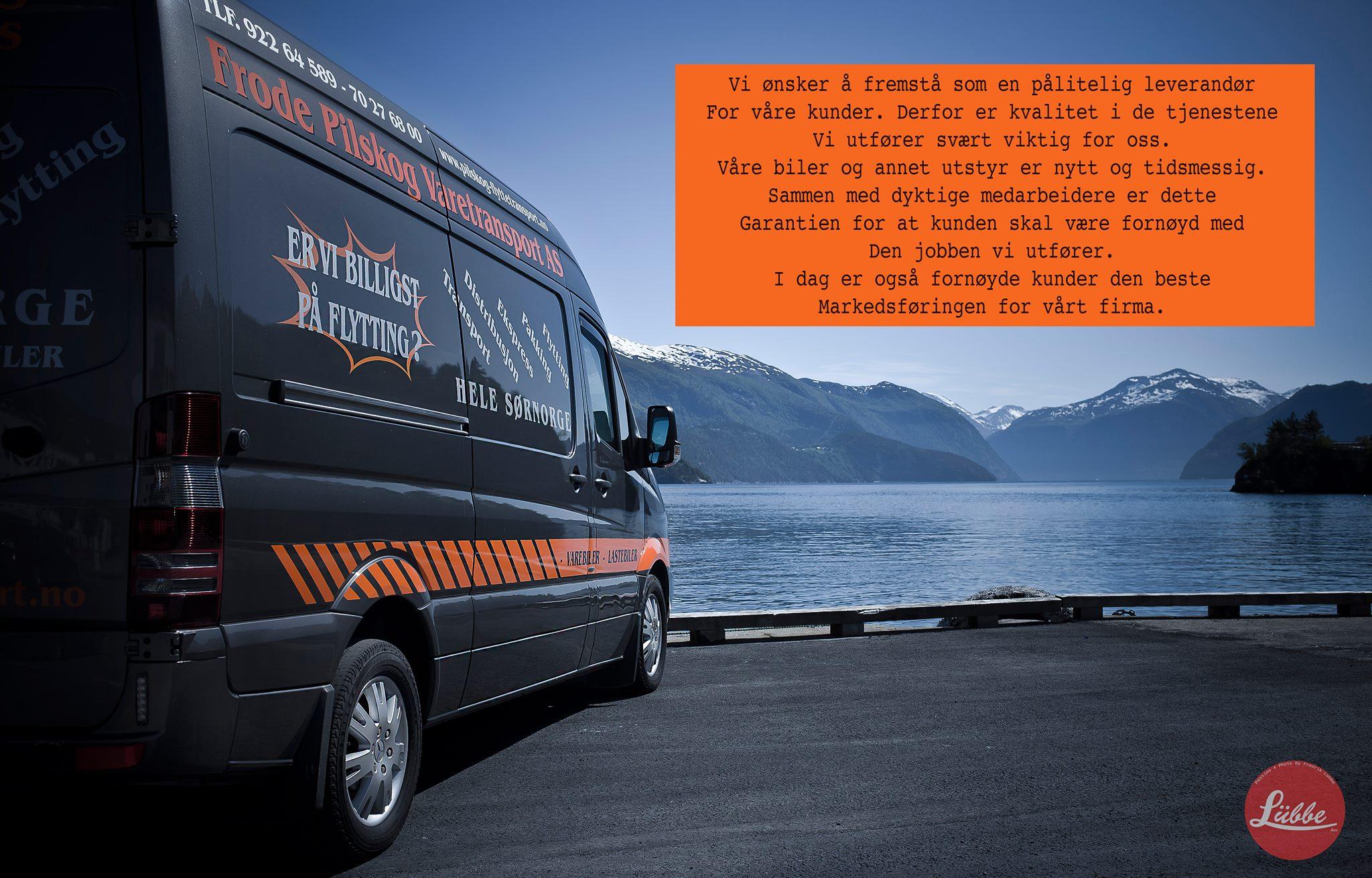 En varebil med logo står ved en vakker fjord med fjell i bakgrunnen og klar blå himmel.
