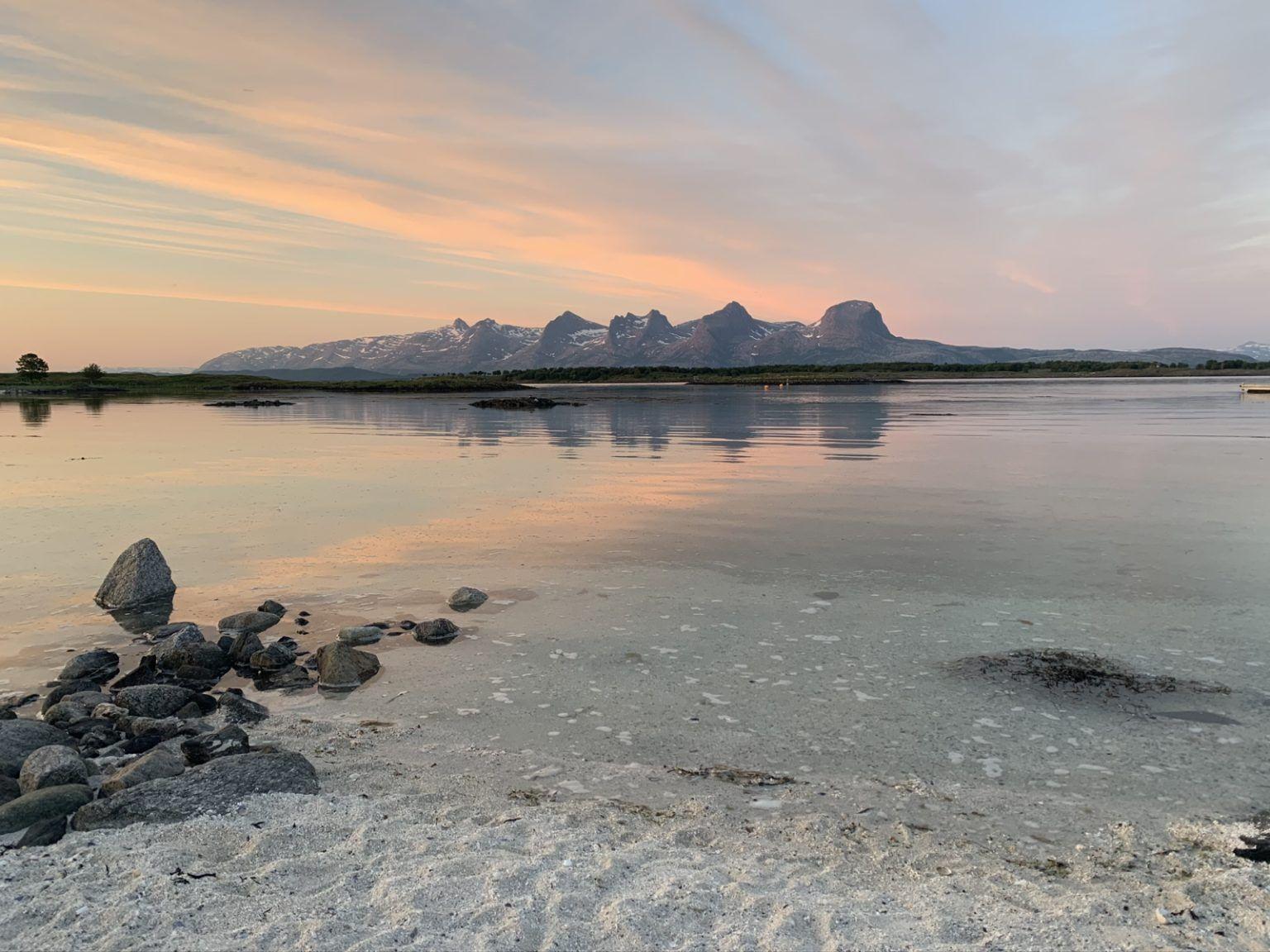 En vakker solnedgang over en rolig innsjø med fjell i bakgrunnen og steiner på stranden.