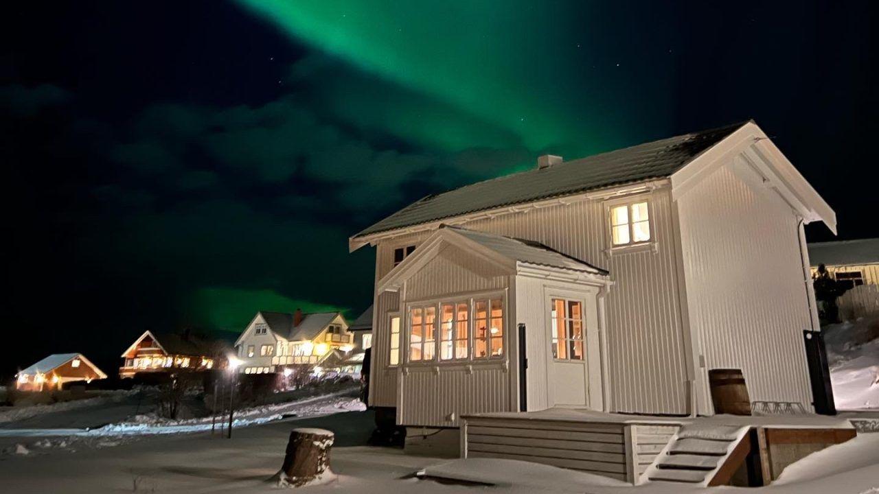 Hvit hytte med lys i vinduene under nordlys på nattehimmelen, omgitt av snødekte landskap.