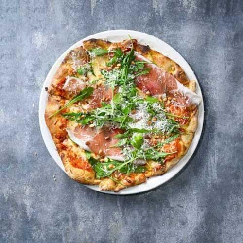 En pizza med skinke, ruccola og revet ost på en hvit tallerken, plassert på en grå overflate.