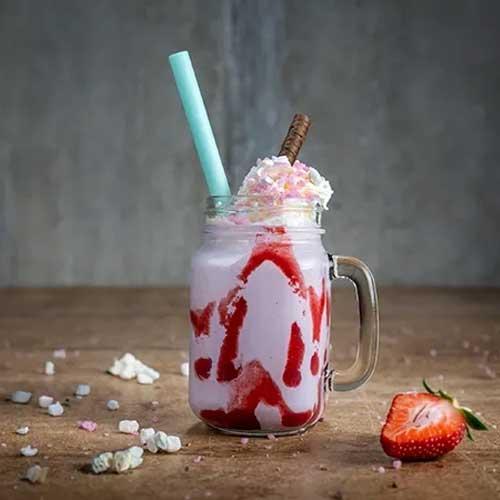 En rosa milkshake med jordbærsaus, krem og en blå sugerør i et glass, med en jordbær ved siden av.