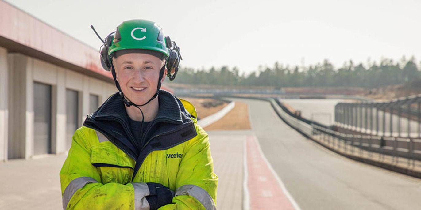 En smilende ung mann i grønn hjelm og refleksjakke står på en racerbane.