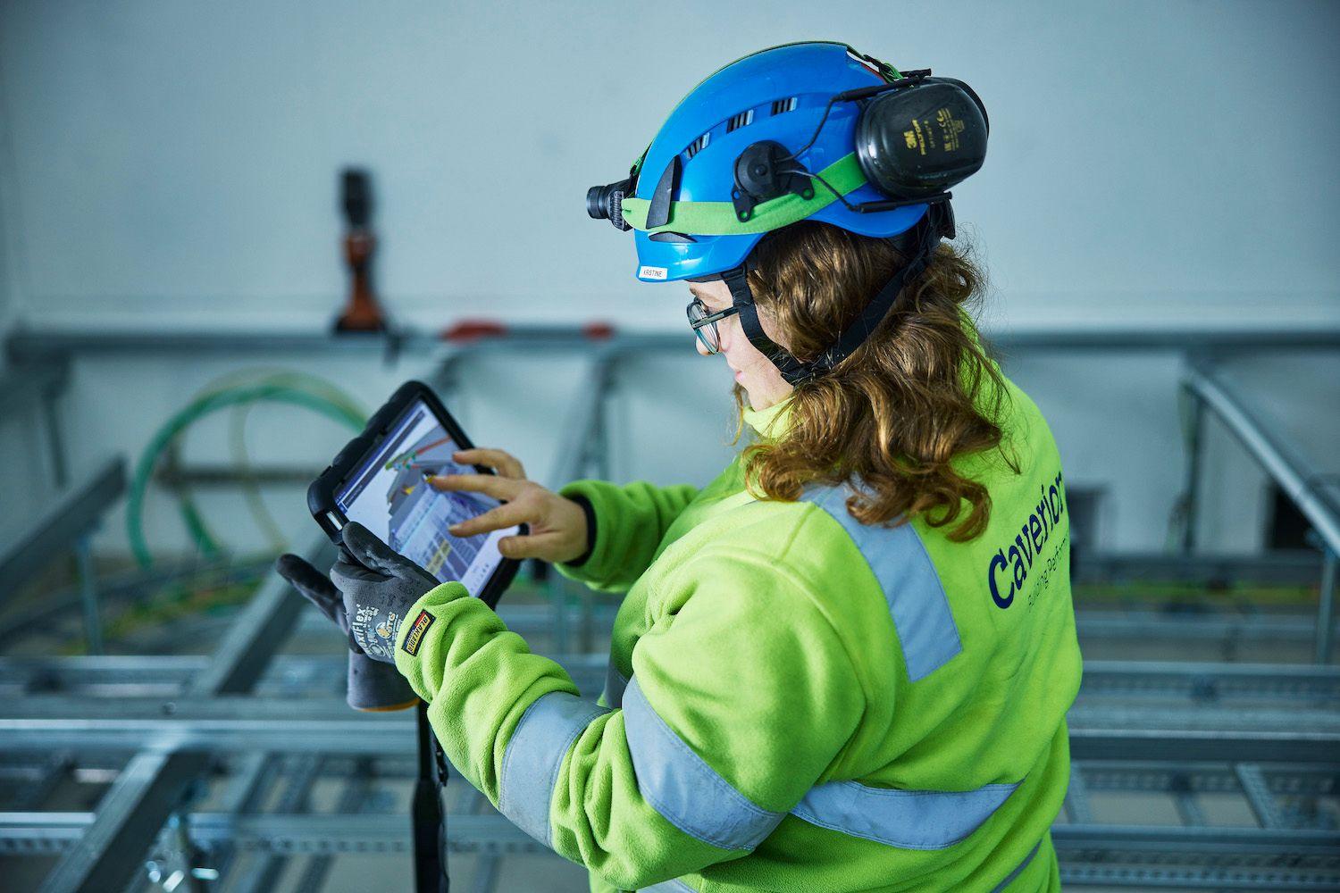 Kvinne med hjelm og verneutstyr bruker nettbrett på byggeplass med metallrammer i bakgrunnen.
