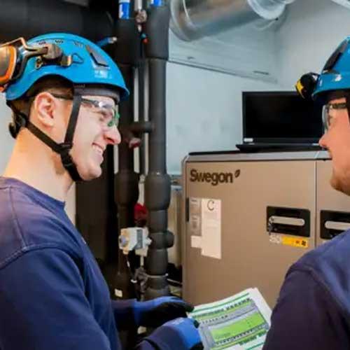 To personer med hjelm og vernebriller som diskuterer en rapport ved en maskin i et industrielt miljø.