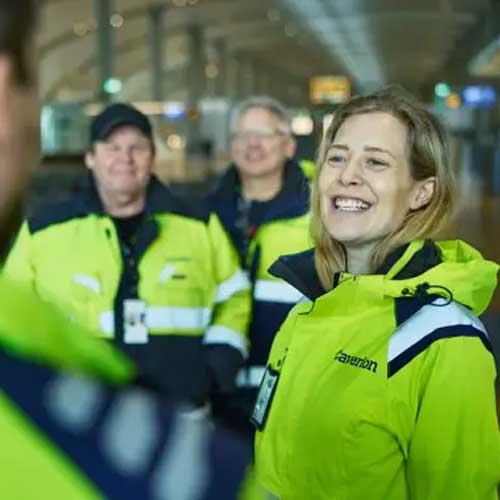 En smilende kvinne i gul jakke står foran to menn i lignende jakker på en flyplass.