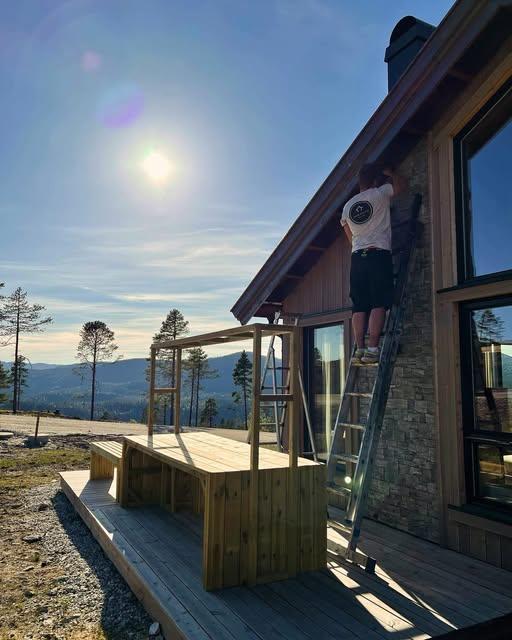 En person står på en stige og jobber med fasaden på en hytte under en solfylt himmel.