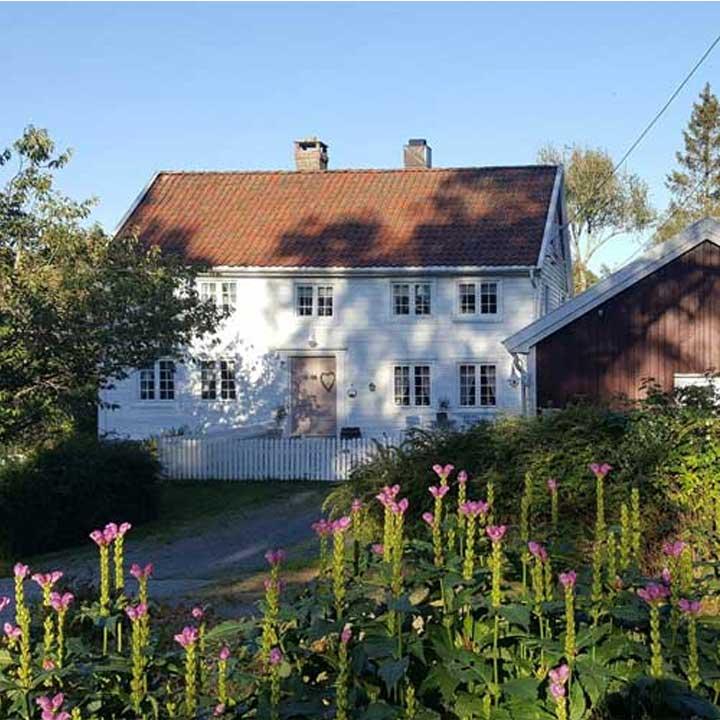 Et hvitt hus med rød takstein, omkranset av blomster og trær, med en liten rød bygning i bakgrunnen.