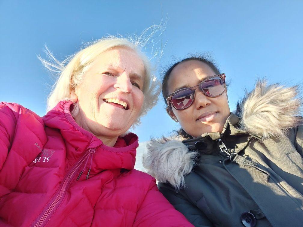 To personer som smiler mot kameraet, med solbriller og jakker, under en klar blå himmel.