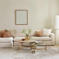 Lys stue med en beige sofa, puter i brunt, et rundt bord og en stor lampe i hjørnet.