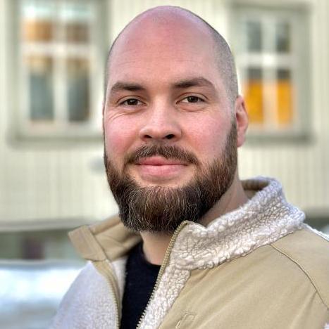 En mann med skjegg og kort hår smiler mot kameraet, iført en beige jakke, med bygninger i bakgrunnen.