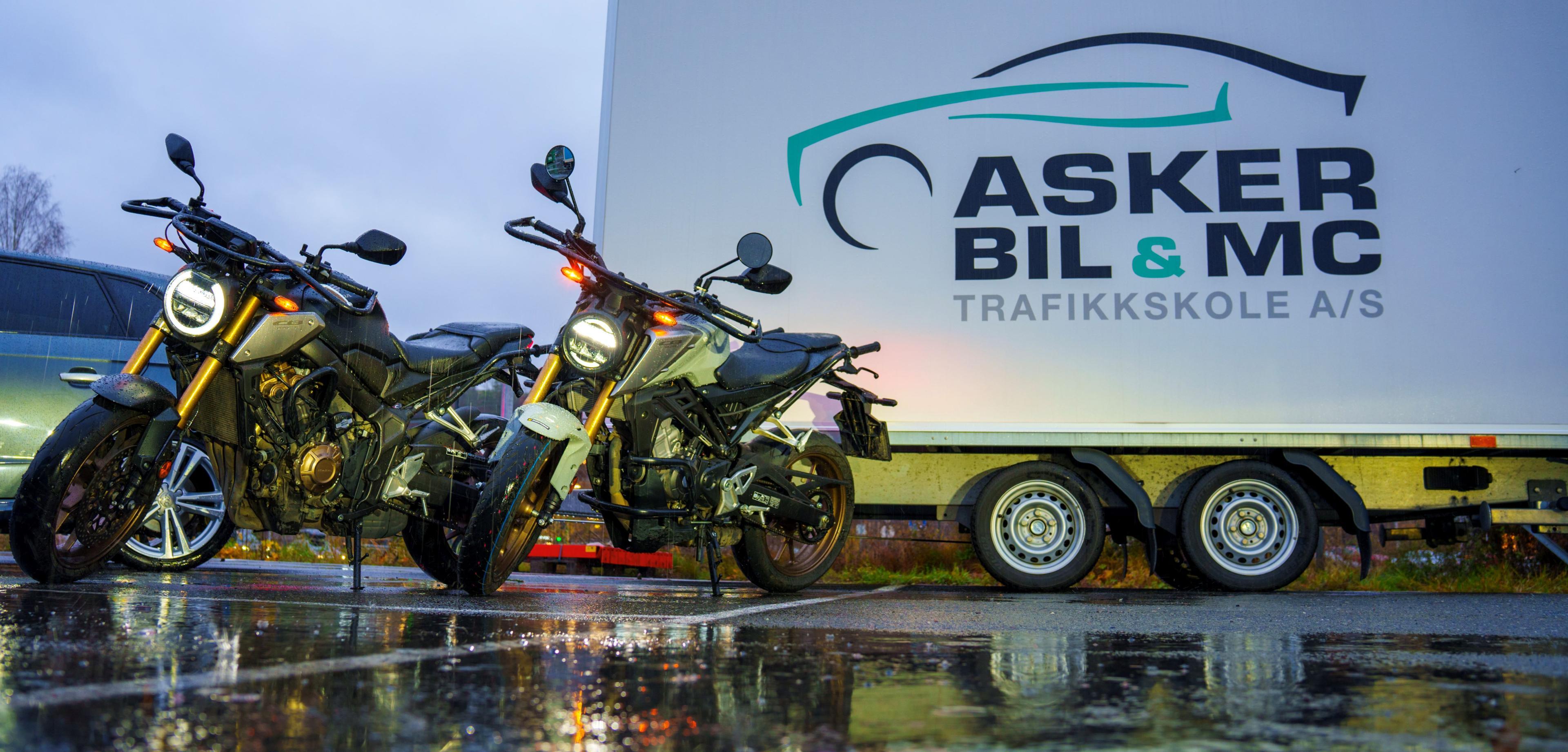 To motorsykler parkert foran en stor trailer med Asker Bil & MC Trafikkskole-logo. Våt asfalt reflekterer lys.