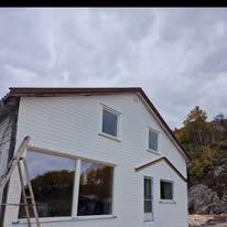 Et nyoppusset hus med hvit kledning, store vinduer og en stige foran, omgitt av natur og grå himmel.