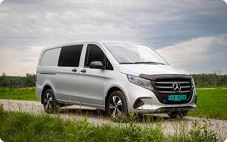 En sølvfarget Mercedes varebil parkert på en grusvei med grønt landskap i bakgrunnen.