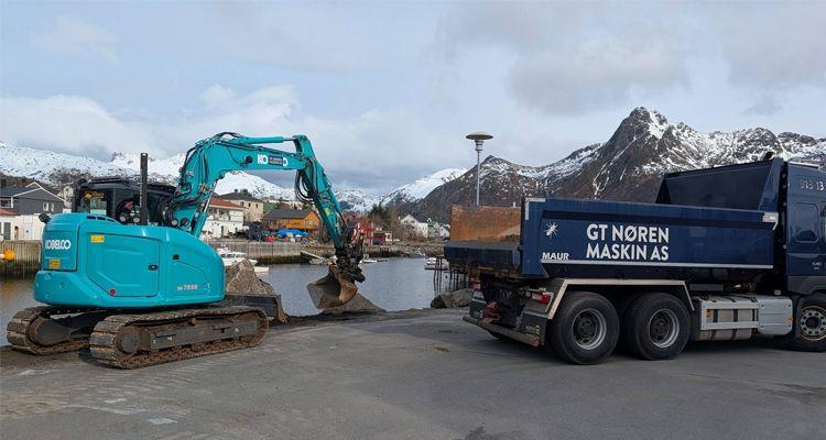 Gravemaskin i arbeid ved havnen, med en lastebil fra CT Noren Maskin AS i bakgrunnen og fjell i horisonten.