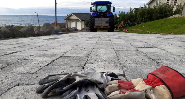 En blå traktor står på en gruslagt gårdsplass med arbeidshansker i forgrunnen og havet i bakgrunnen.