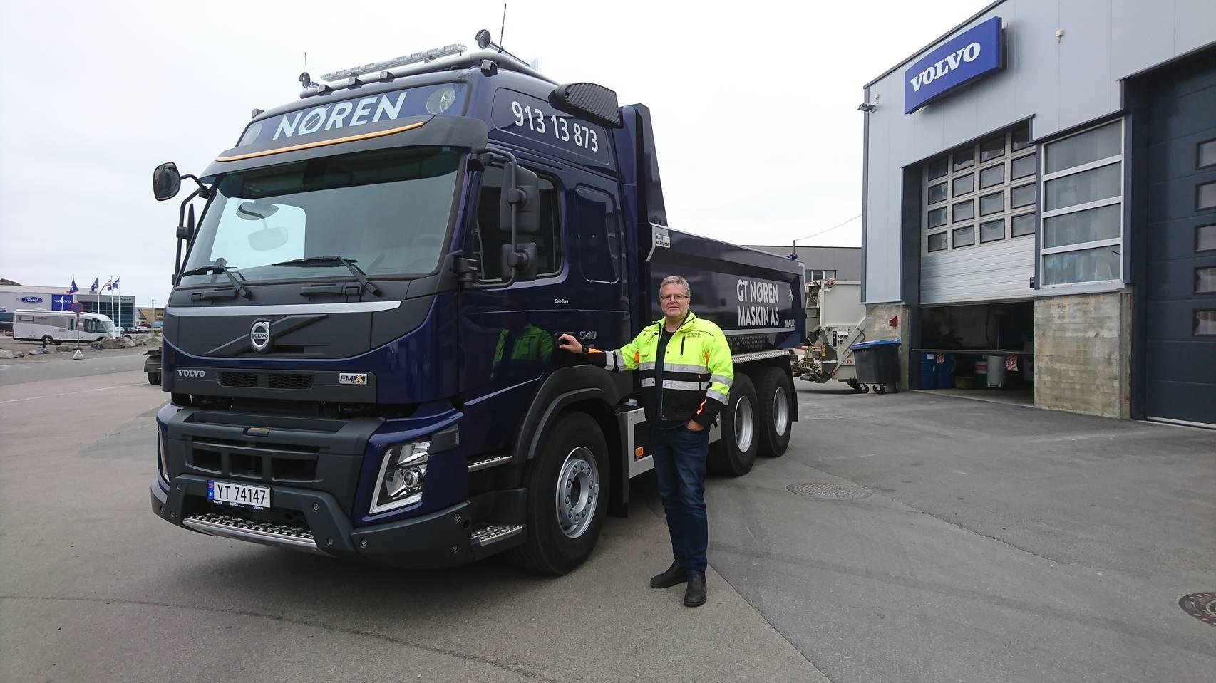 En sjåfør står ved siden av en blå Volvo lastebil med Noren-logo, foran en verkstedbygning.