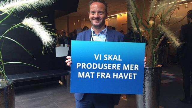 Mann holder et blått skilt med teksten "Vi skal produsere mer mat fra havet" på en konferanse.