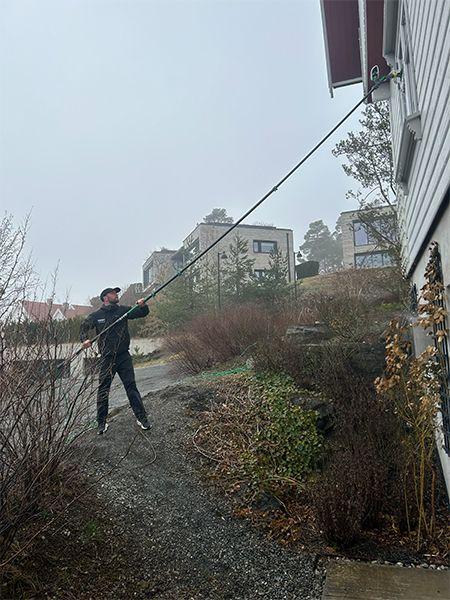 En person bruker en lang stang for å nå opp til et vindu på en bygning i regnvær. Boliger i bakgrunnen.