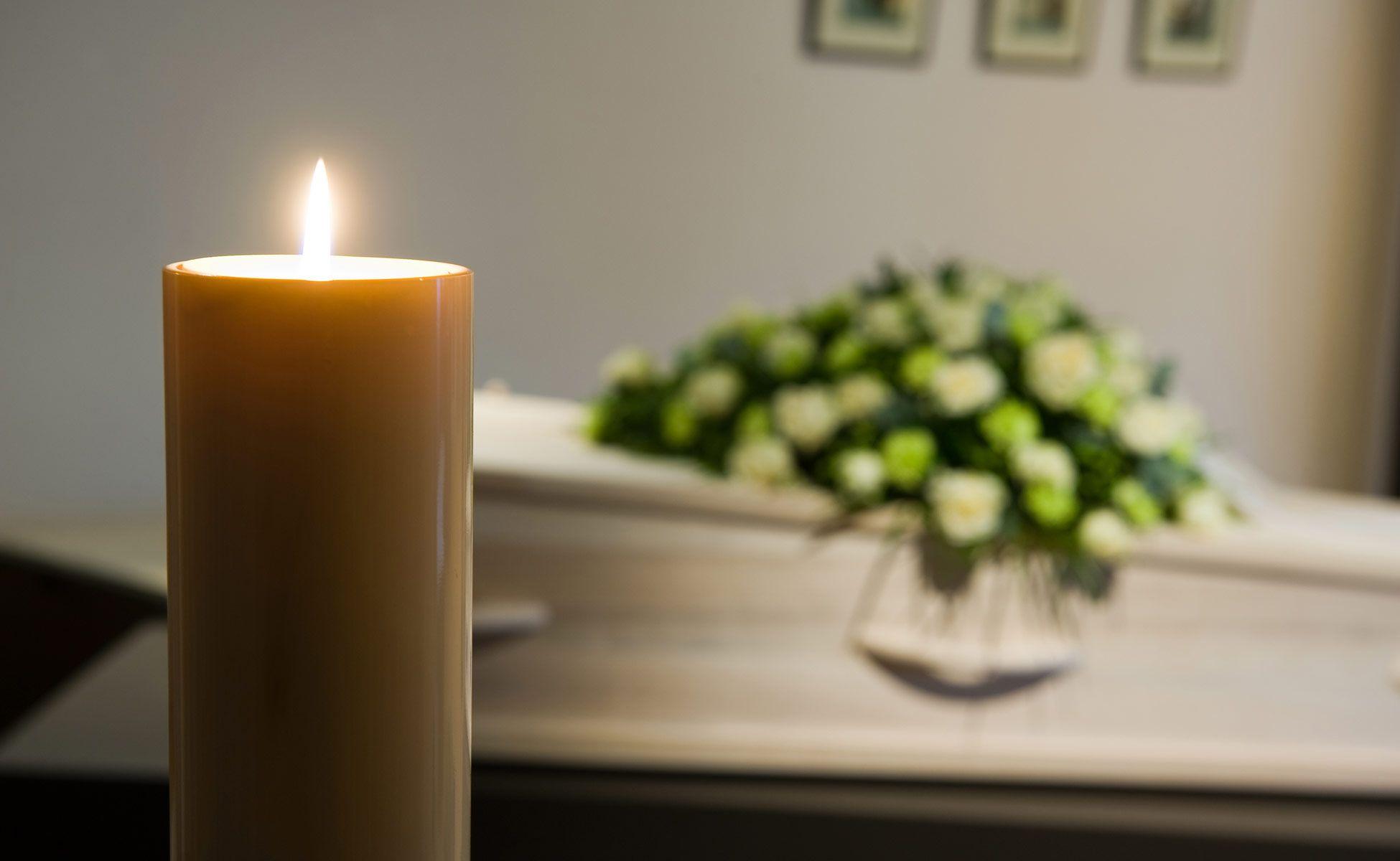 Et tent lys foran en kiste med blomsteroppsats i bakgrunnen, som skaper en stille og respektfull atmosfære.