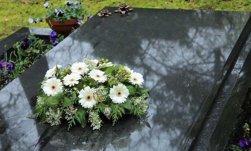 Blomsterdekorasjon med hvite gerberaer og grønt på en gravstein i en hage med blomster.