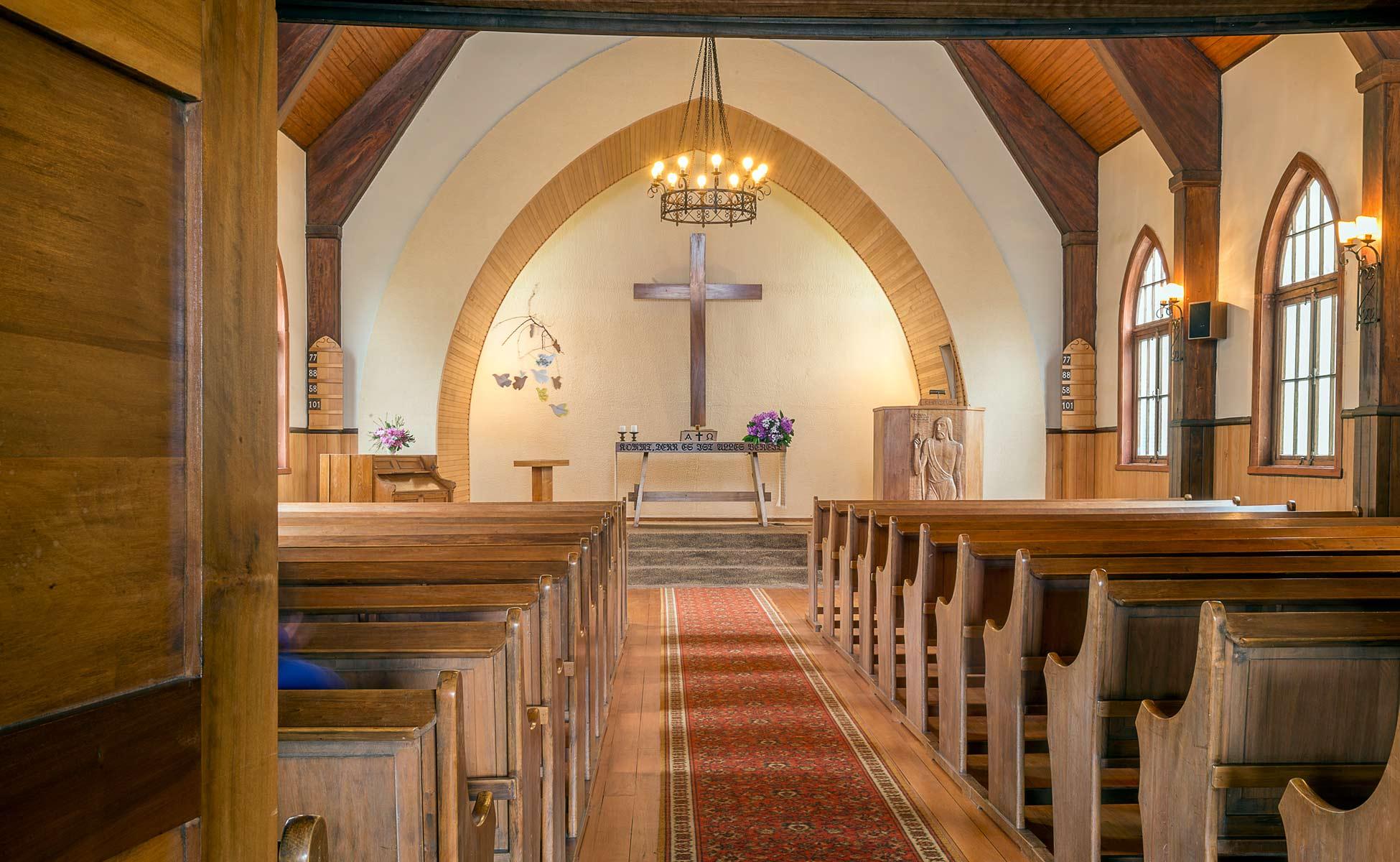 Interiør av en kirke med trebenker, alter, kors og blomsterdekorasjoner i et lyst rom.