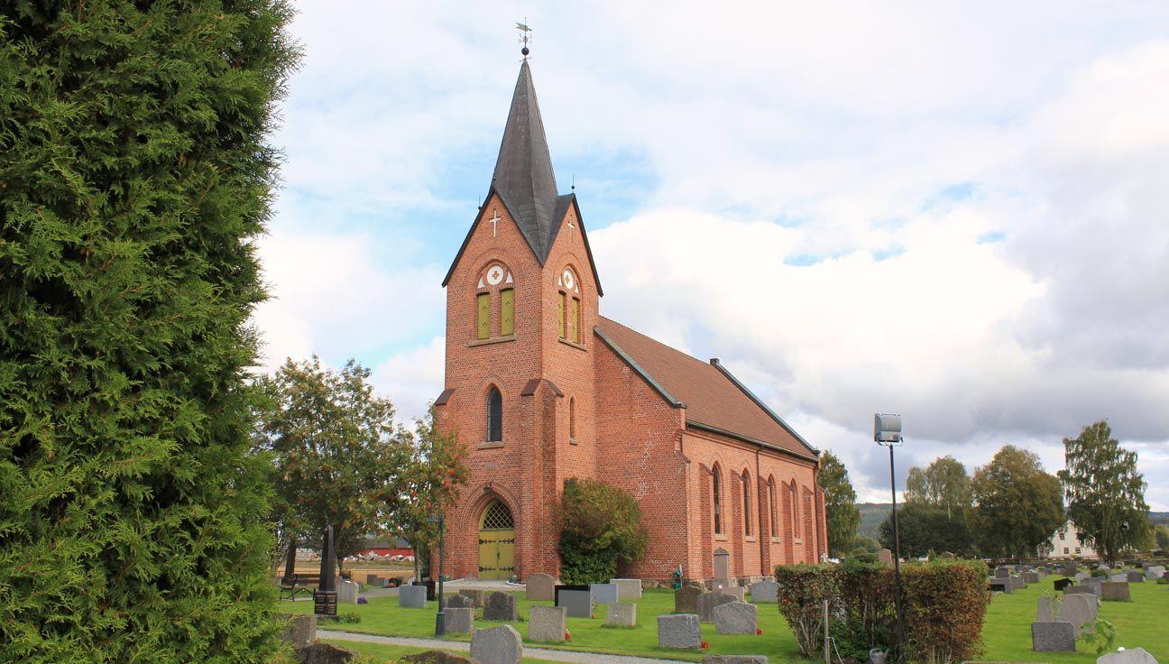 Rød mursteinkirke med spisstårn, omgitt av gravsteiner og trær på en overskyet dag.