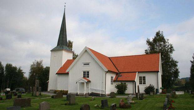 En hvit kirke med spisstårn og rød takstein, omgitt av gravsteiner og trær.