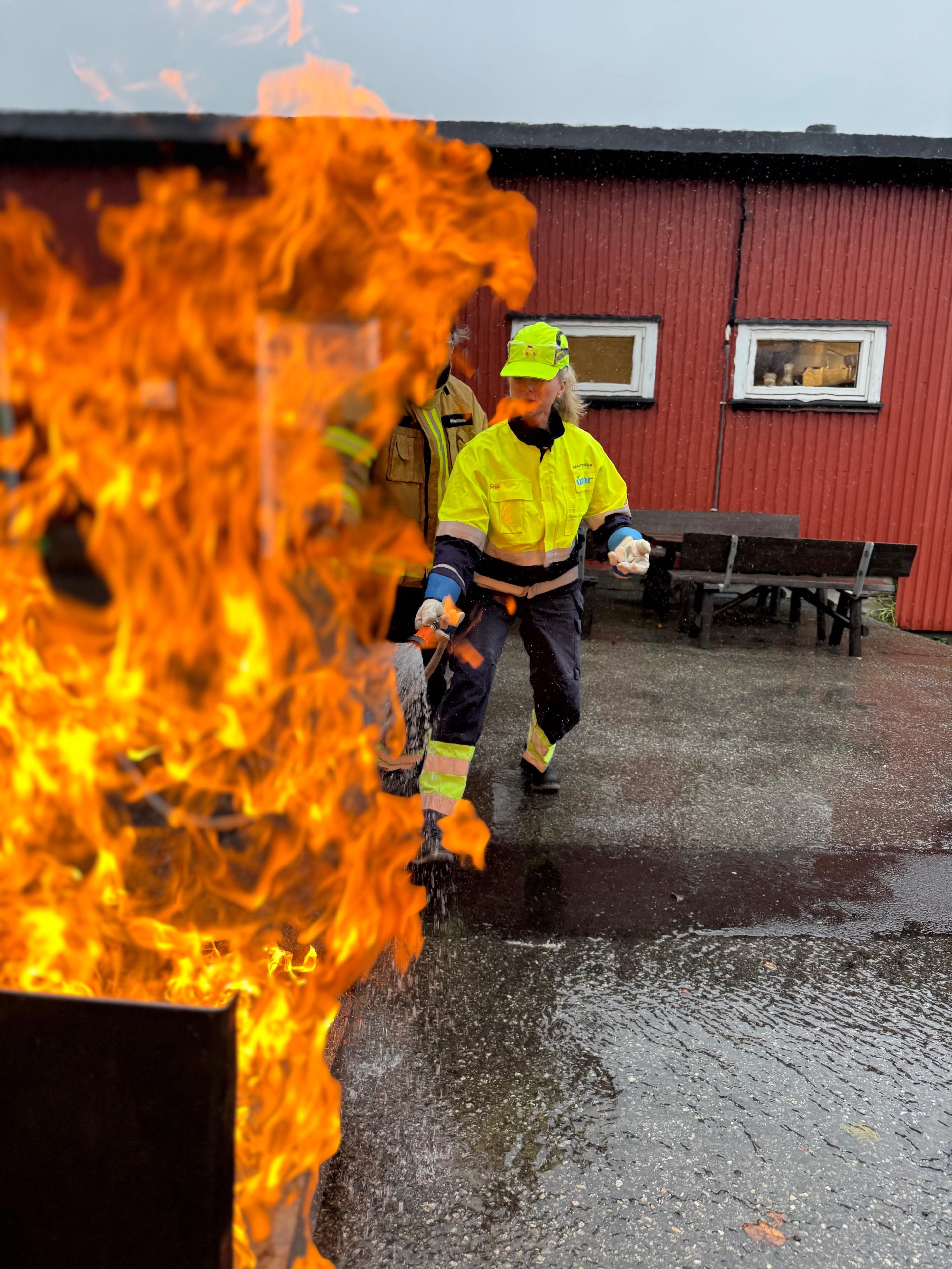 Brannmann i sikkerhetsutstyr slukker brann ved en bygning, med flammer i forgrunnen.