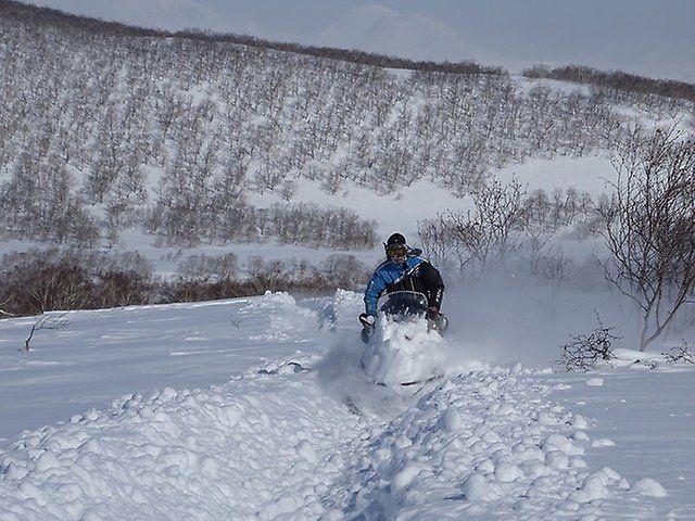 En person kjører snøskuter gjennom snødekt landskap med fjell i bakgrunnen.