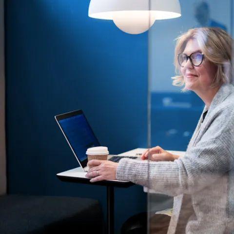 Kvinne med briller sitter ved et bord med laptop og kaffekopp i moderne kontormiljø.