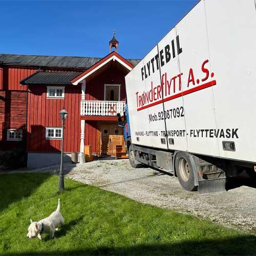 En flyttebil fra Trønderflytt A.S. står parkert foran et rødt hus med en hund som går på plenen.