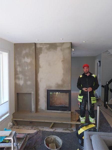 En håndverker står foran en uferdig peis i et rom med grå vegger og moderne interiør.