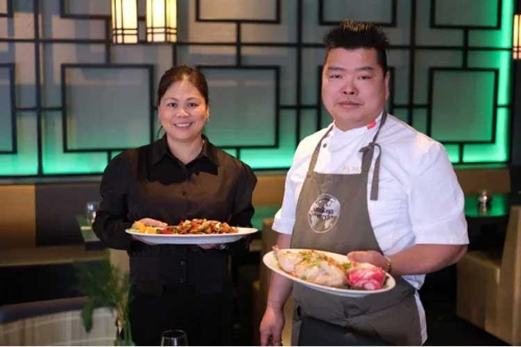 To personer i en restaurant holder tallerkener med mat, en kvinne i svart skjorte og en mann i kokkejakke.