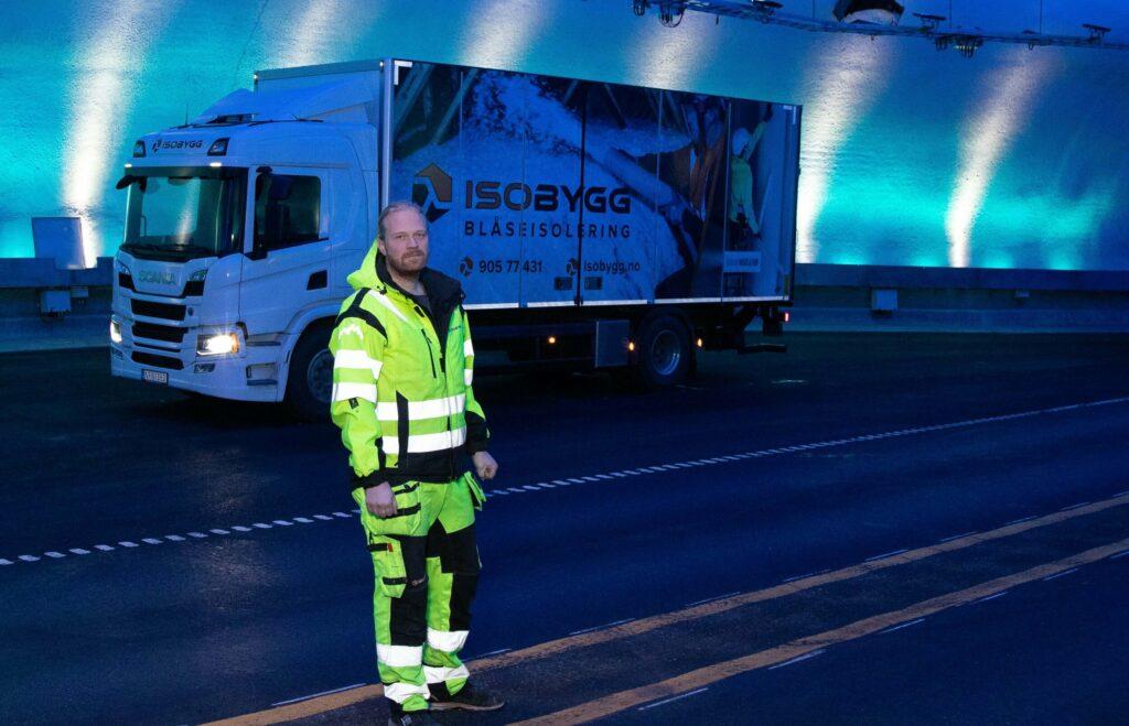En mann i refleksjakke står foran en lastebil i en opplyst tunnel. Lastebilen har reklame for Isolbygg.