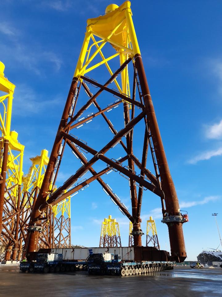 Store gule vindturbinfundamenter står på en havn under en klar blå himmel.