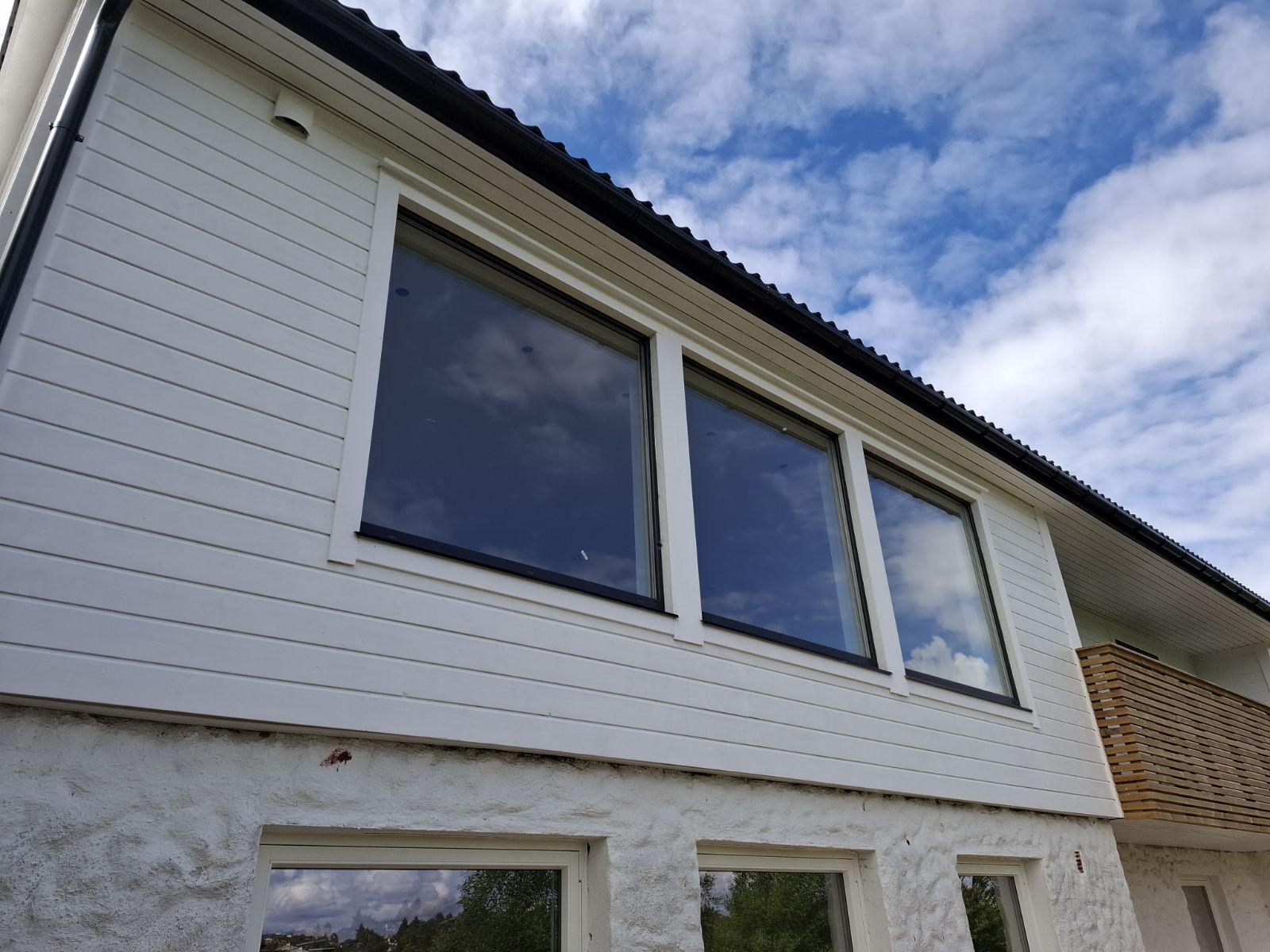 Fasade av et hus med store vinduer og hvit panel, under en blå himmel med skyer.