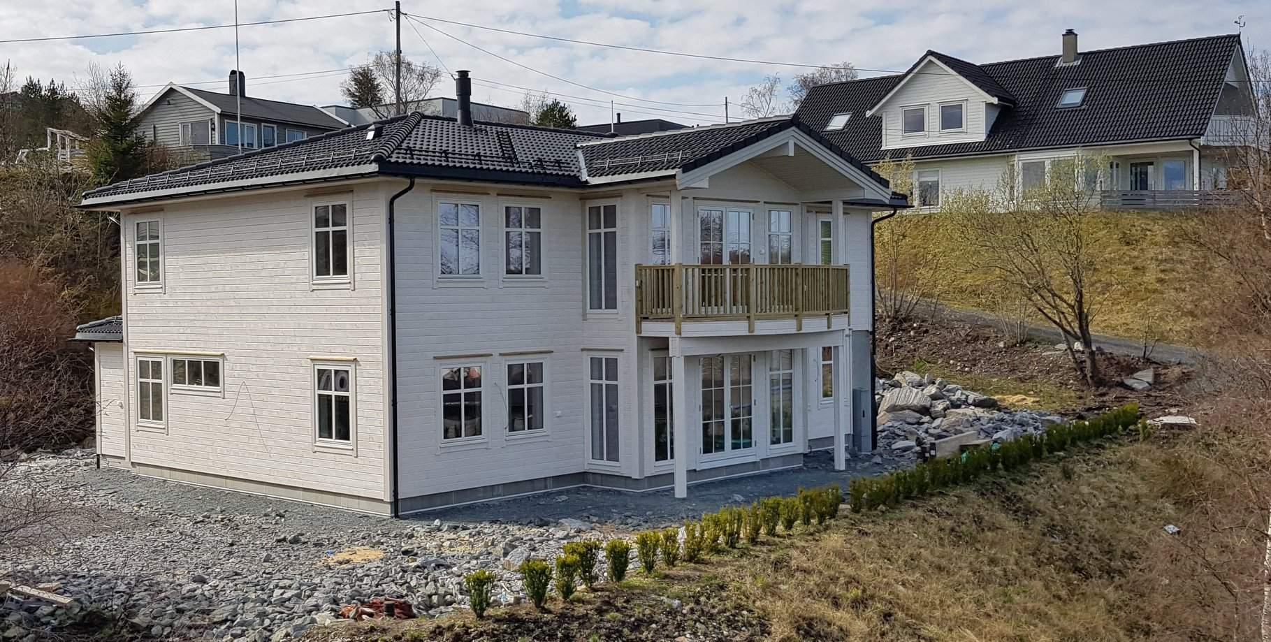 Moderne, hvitt hus med balkong og steinlagt hage, omgitt av grønne områder og andre boliger.