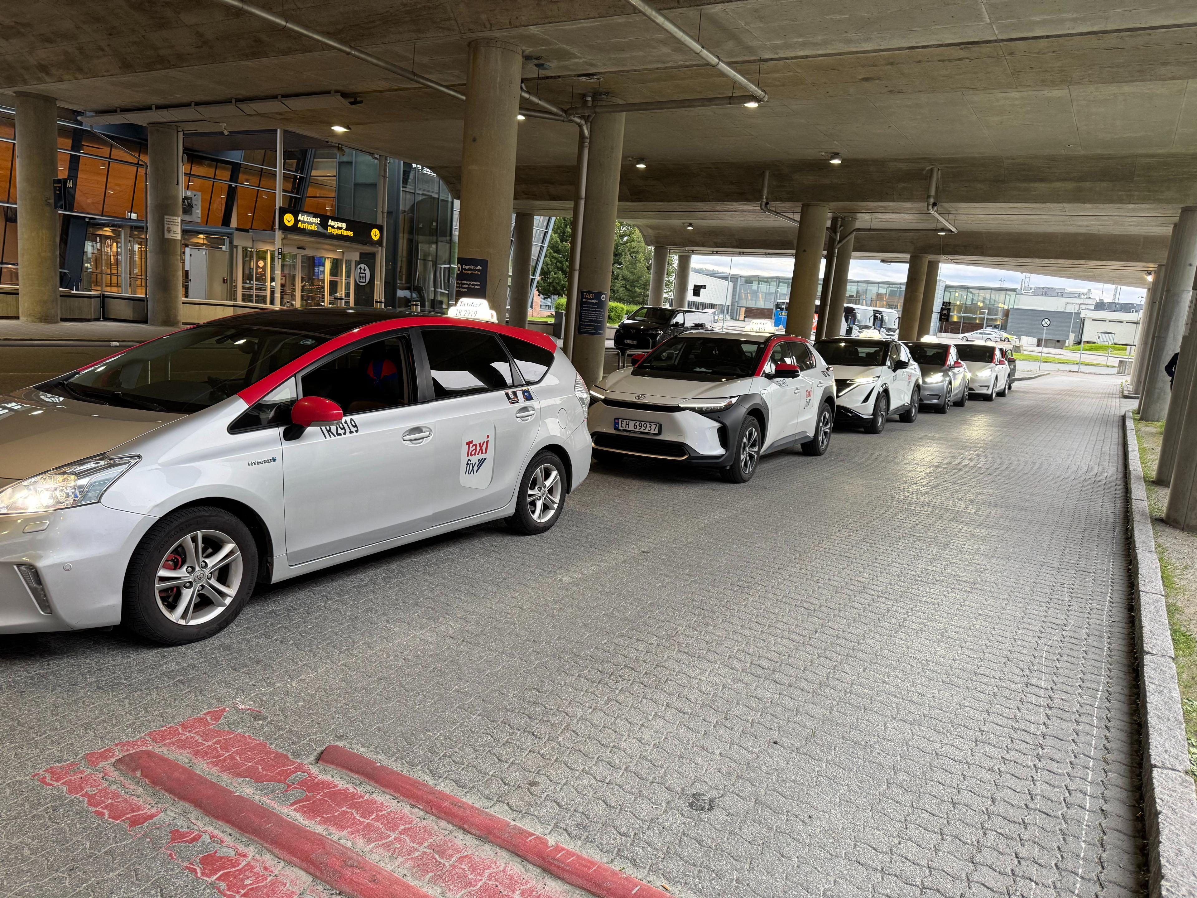 En rekke grå taxier med røde tak parkert under en bygning.